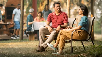 A courting couple sitting together at a social gathering. They enjoy talking with each other while others associate in the background.