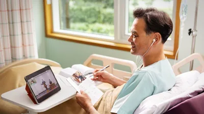 A brother in a hospital bed, attending the “Watchtower” Study via videoconference. He follows along with his own copies of “The Watchtower” and the Bible.