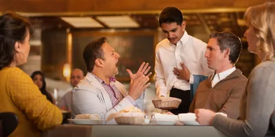 A man dining in a restaurant, berating a waiter who is trying to apologize. Others at the man’s table appear uncomfortable.