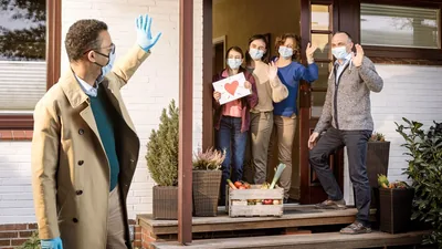 A brother delivering a box of food to a family. All are wearing masks and wave from a distance.