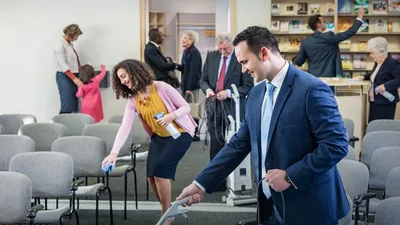 Brothers and sisters joyfully cleaning a Kingdom Hall. In the background, others are putting a donation in the contribution box, conversing, and organizing literature for the congregation.