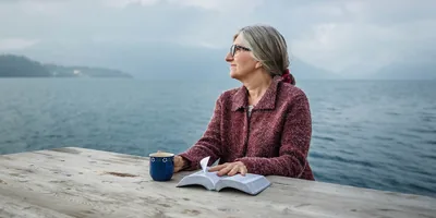 A woman holds an open Bible, sits at a table, and stares into the distance