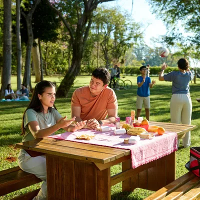 A family enjoying time together at a park. The father listens attentively to their teenage daughter as she expresses herself while the mother plays ball with their young son.