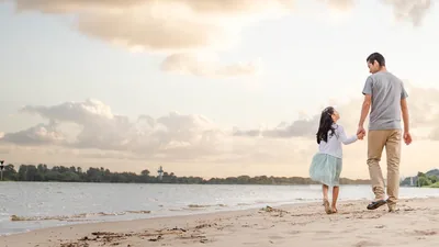 A father holding his young daughter’s hand as they walk along the shore.