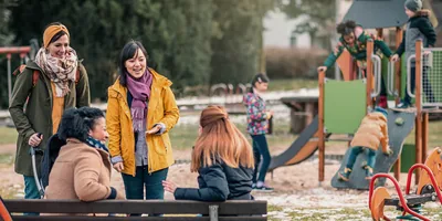 Four women of different racial backgrounds talking and laughing as their children play together at a playground.