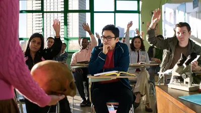 A young brother in school, contemplating as his teacher holds a misshapen human skull in front of the class. The majority of his classmates have their hands raised.