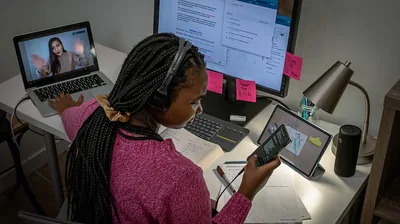 A teenage girl multitasking. She types a text message on her smartphone, video chats with a friend, and uses a computer and textbook to study.