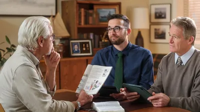 The older man from the previous image studying the “Enjoy Life Forever!” book with two brothers. A display of military medals is on a nearby shelf.
