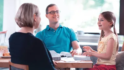 A father conducting family worship with his wife and young daughter. He smiles as his daughter expresses herself enthusiastically.