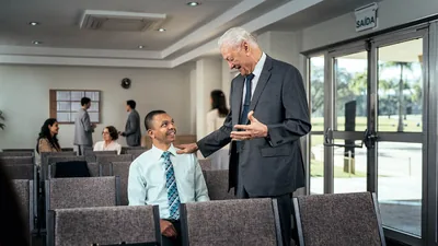 An elder speaking warmly to a younger brother who is sitting alone at the Kingdom Hall.