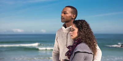A grieving couple on the beach hold each other and look into the distance