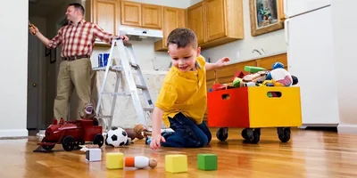 A little boy picks up his toys while his father paints a kitchen wall