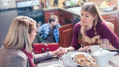 A couple helping a sister in need. The wife listens empathetically to the sister while her husband repairs the sister’s kitchen sink.