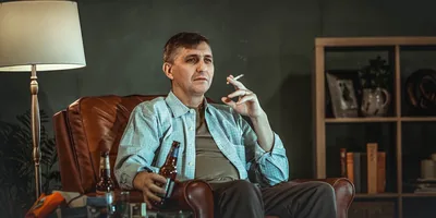 A man sitting in his living room, holding a cigarette in one hand and an opened beer bottle in his other hand.