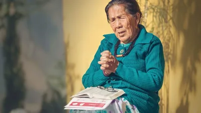 An elderly sister praying intensely. On her lap are a Bible and a list from jw.org of brothers in prison.