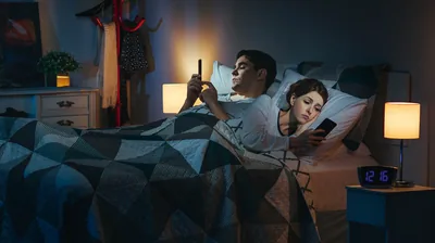 A husband and wife each using a smartphone while lying in bed late at night.