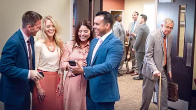 At a Kingdom Hall, two couples laughing while looking at a cell phone. In the background, three young brothers talking and enjoying themselves. An older brother looking disappointed that he is not invited to join any of these conversations.