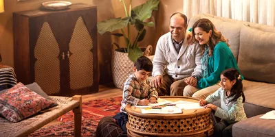 A family happily spending time together. The parents sit on a sofa holding hands while the children draw pictures at the coffee table.
