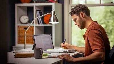 A young brother using the Bible, a tablet, and a notebook to do his personal study. He writes a note in his Bible.