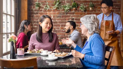 A young sister listening to an older sister’s advice while they eat a meal together at a restaurant.
