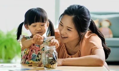 A mother helping her young daughter put money into a jar.