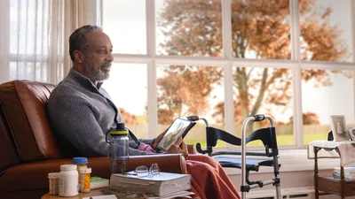 The elderly brother sitting in his home in the autumn season and thinking about the picture of Paradise displayed on his tablet. Beside him is a walker and several bottles of medication.