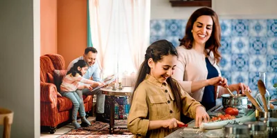 A family happily spending time together. The mother prepares a meal with one of the daughters in the kitchen. The father looks at a photo album with the other daughter in the living room.