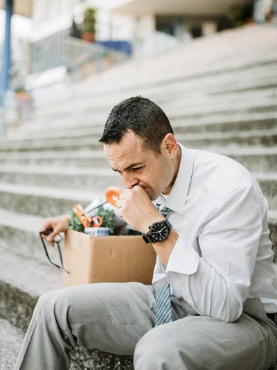 An upset businessman sitting on the steps of a building. A box of personal office items is beside him.