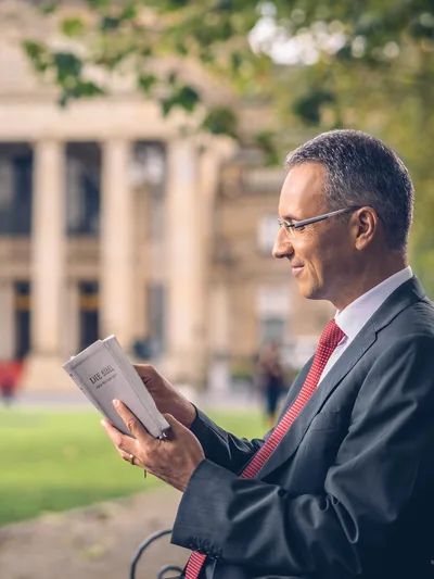 A man reading a Bible while sitting on a bench in front of a government building.