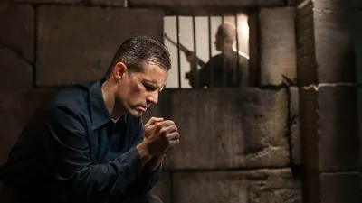 A brother in a prison cell, praying intensely. Outside the cell, a prison guard stands watch, holding a gun.