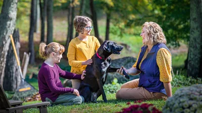 A mother and her two young daughters conversing outdoors and petting their dog.