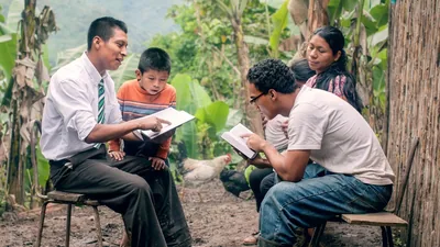 A brother reading a scripture to a family outside their modest home. The father follows along in his Bible while the mother and young son listen intently.