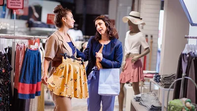 Two sisters shopping at a clothing store. One of the sisters suggesting to the other sister that the short skirt she is holding may be inappropriate.