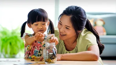 A young girl dropping coins into a jar as her mother joyfully observes.