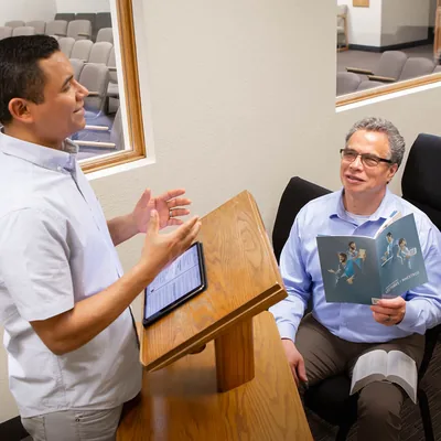 An elder listening to a brother practice a talk. The elder holds an open “Teaching” brochure.