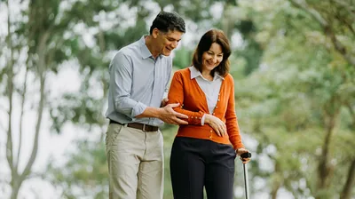 The couple who earlier were in the hospital, walking together in a park. The husband gently assists his wife to use a cane.