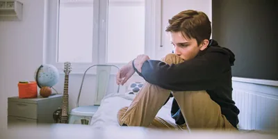 A teenage boy sits on his bed looking depressed