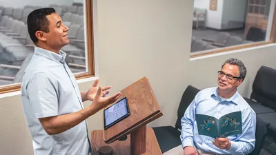 An elder listening to a brother practice a talk. The elder holds an open “Teaching” brochure.