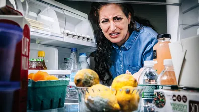 A woman looking with disgust at rotten food in a refrigerator.