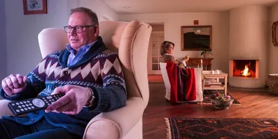 A husband watches television in one area while his wife does needlework in another